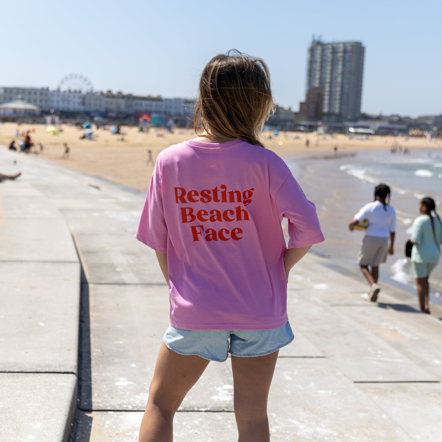 Person wearing a pink t-shirt with 'Resting Beach Face' text at a beachside promenade.