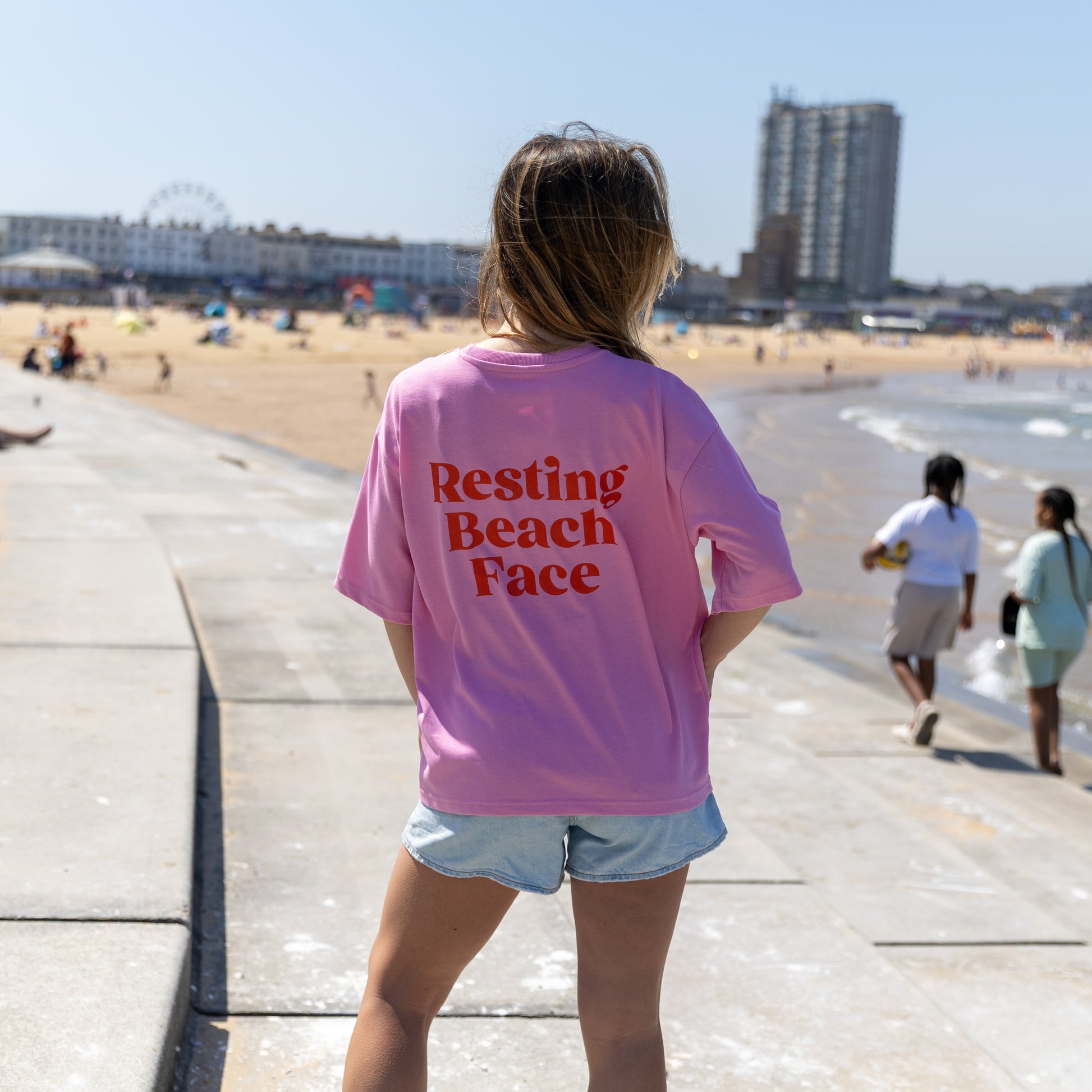 Person wearing a pink t-shirt with 'Resting Beach Face' text at a beachside promenade.
